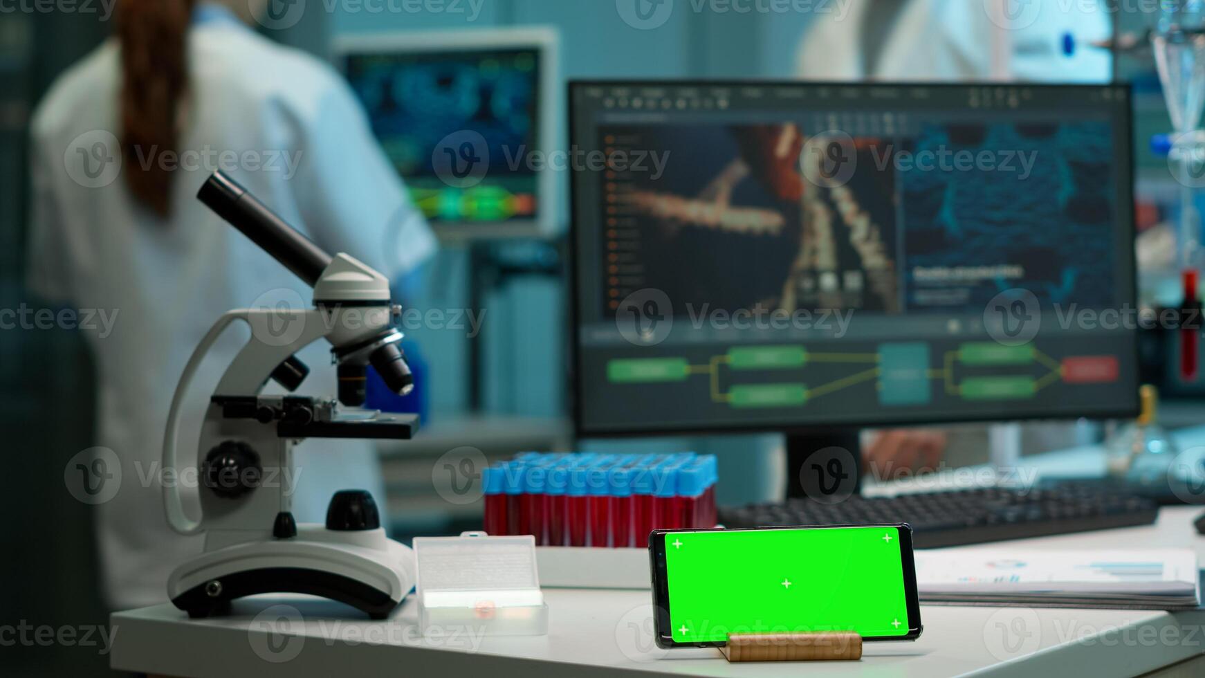 Microbiologist bringing blood samples in modern lab putting them near smartphone working with green chroma key screen placed on desk. Team of biotechnology scientists developing drugs in background photo
