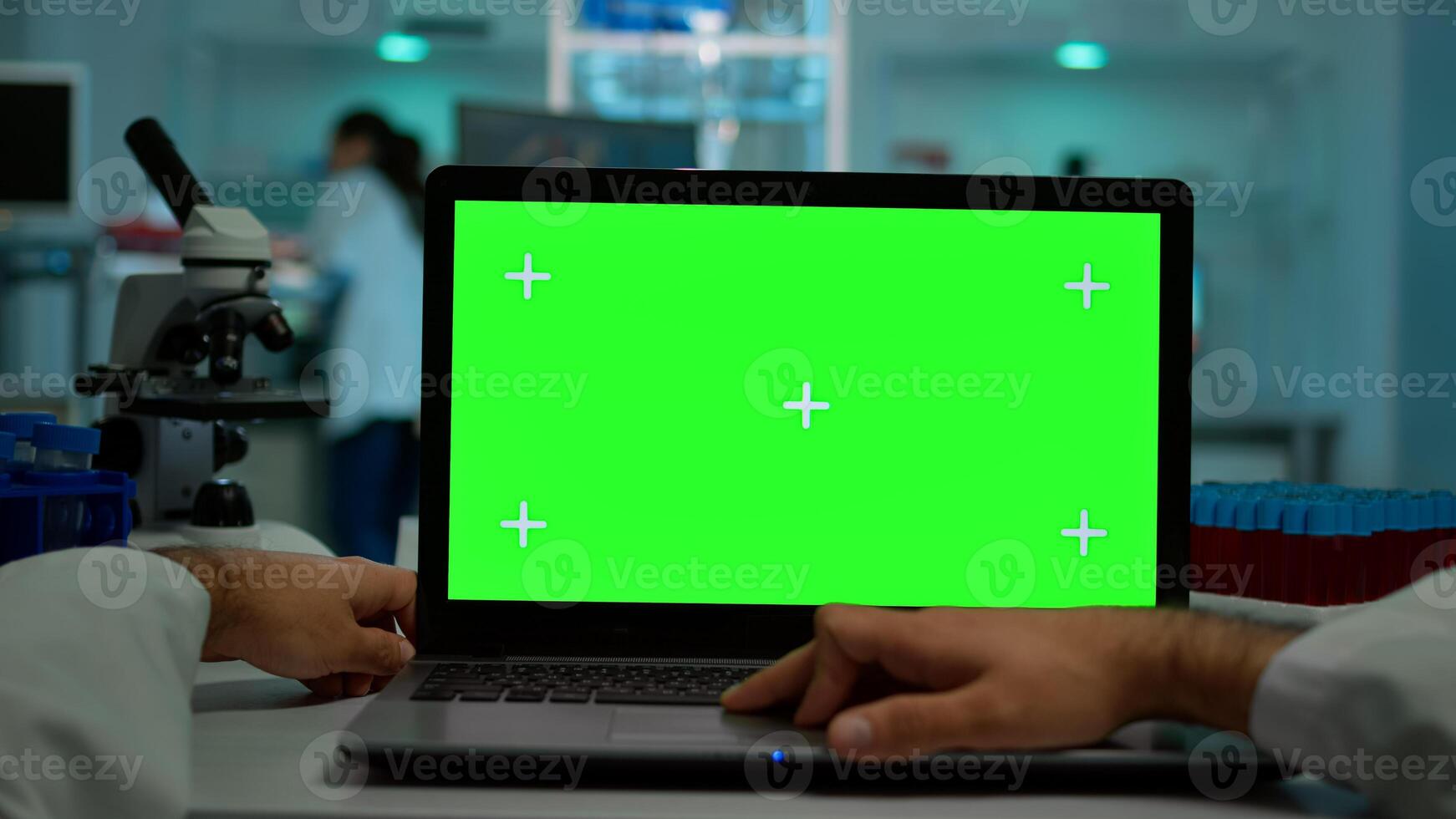 Pov shot of man scientist sitting at desk working on laptop with mock-up green screen, isolated display. In background lab researcher analysing vaccine developent examining samples photo