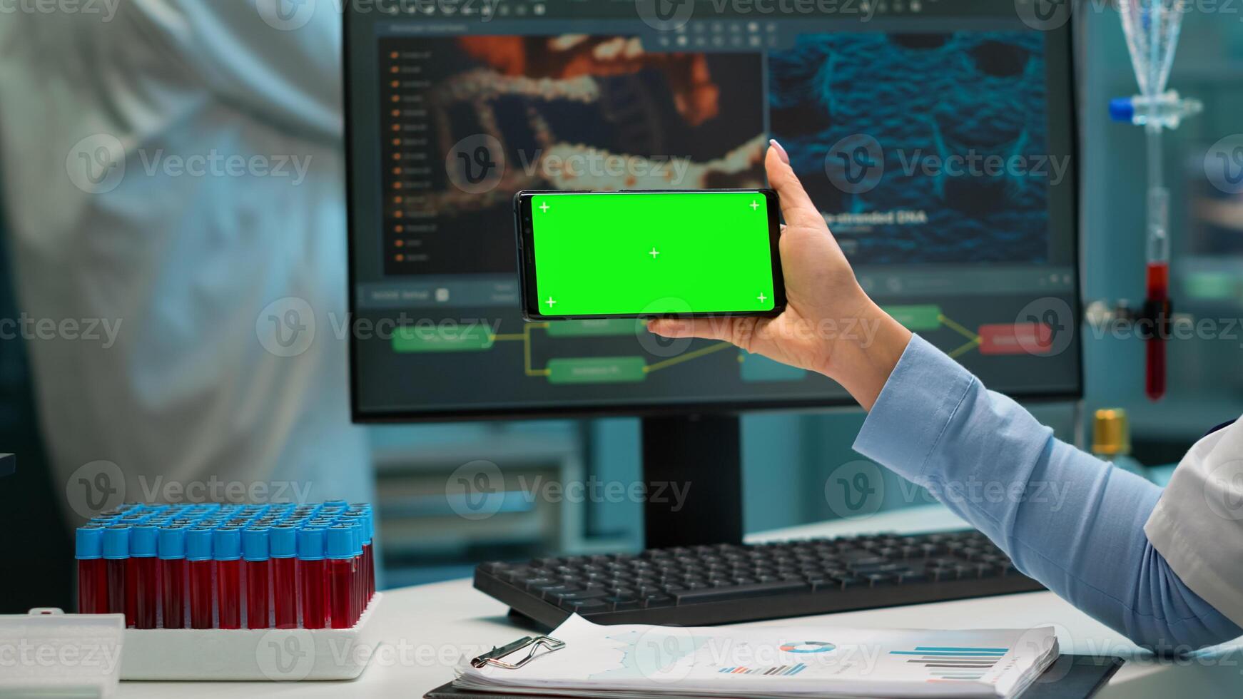Close up of scientist woman holding smartphone with green mockup in modern equipped lab. Team of microbiologists doing vaccine research writing on device with chroma key, isolated display. photo