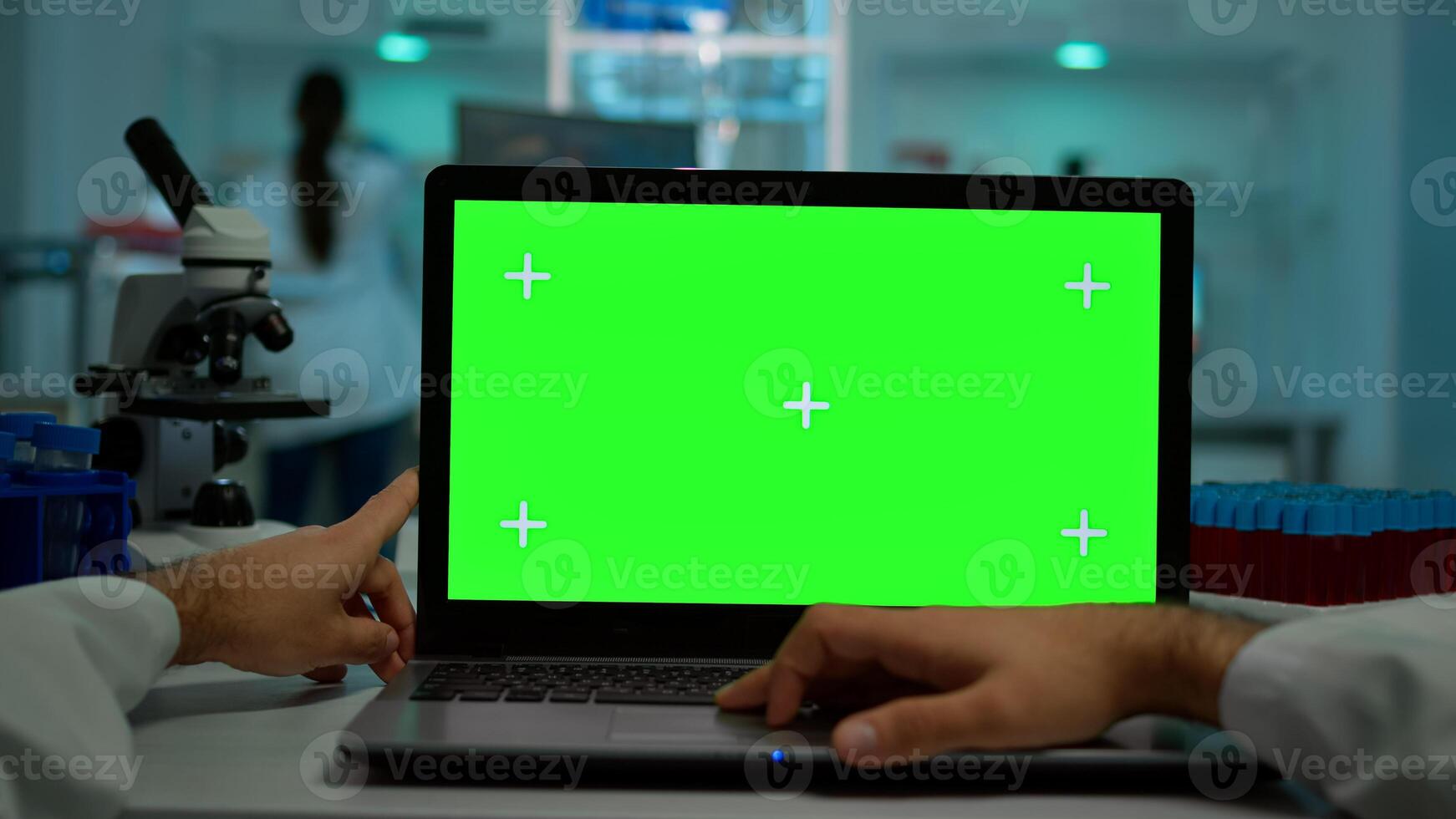 Pov shot of microbiologist typing on laptop with green chroma key display sitting at desk working reading virus symptoms. In background lab researcher analysing vaccine developent examining samples photo