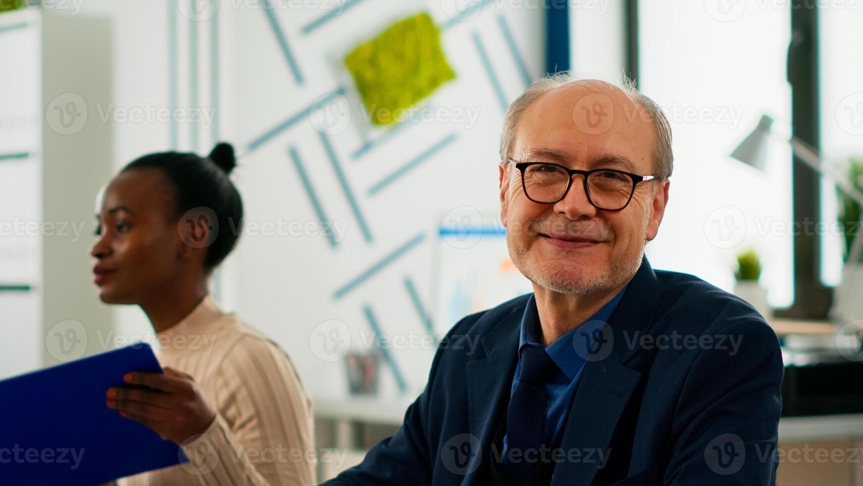 Elderly businessman looking at camera smiling sitting in brainstorming room, preparing for meeting with partners. Manager working in professional start up financial business ready for conference photo