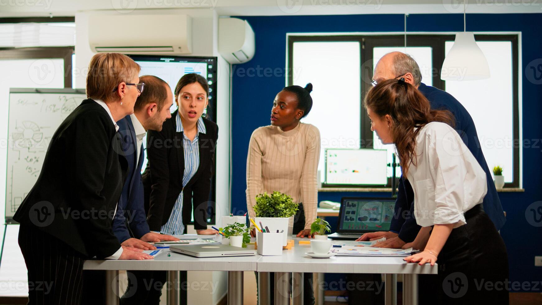 Diverse businesspeople brainstorming while meeting at company office standing at conference desk looking in documents. Colleagues working planning success financial strategy discussing in office photo