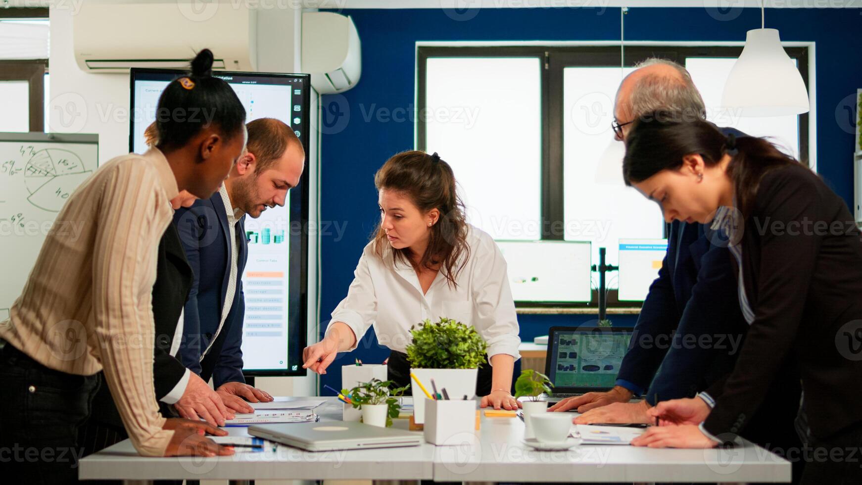 Diverse team of workers talking, looking at documents and analyzing graphs data standing at conference desk in start up company. Professional colleagues team discussing marketing project brainstorming photo
