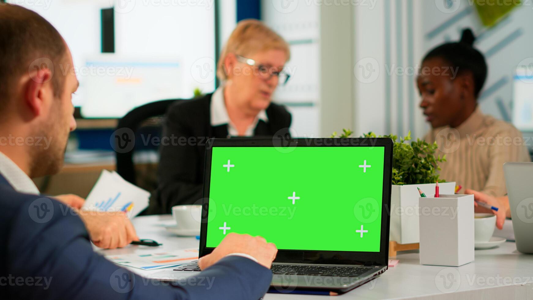 Back view of business man sitting conference desk using laptop with green screen talking with colleague while team working on background. Multiethnic coworkers planning project on chroma key display photo