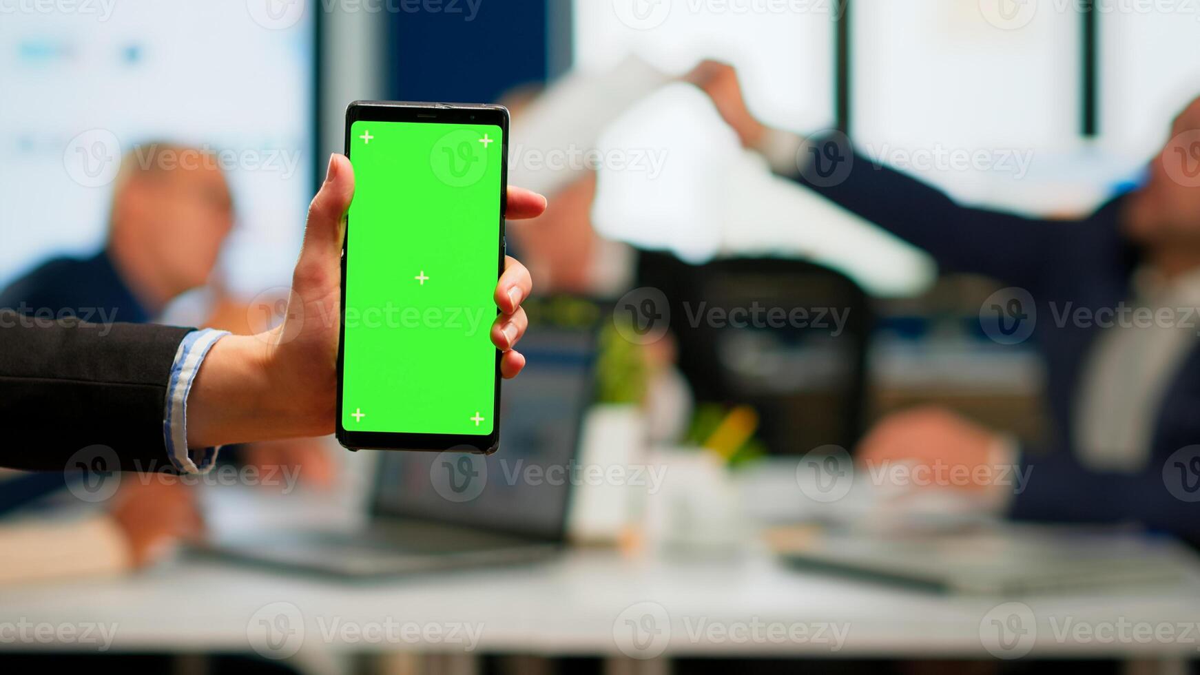 Businesswoman holding smartphone with green screen while diverse team working on background, businesspeople analysing financial statistics, Multiethnic coworkers planning project on chroma key display photo