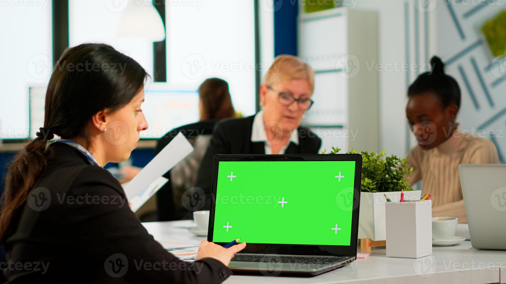 Over the sholder of manager sitting at meeting desk looking at laptop with green screen mockup while diverse team working on background. Multiethnic people planning project on chroma key display photo