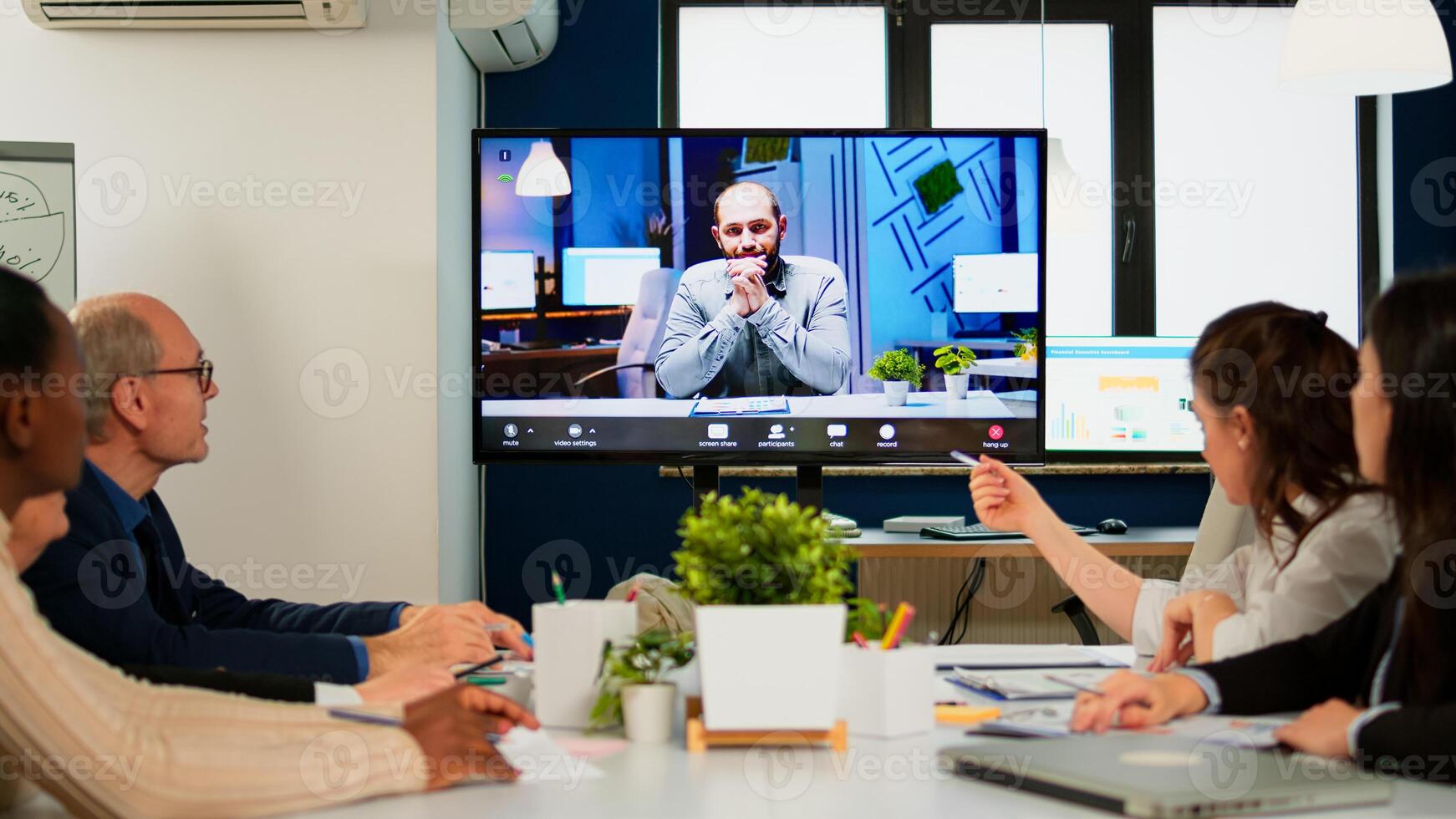 Manager man talking with colleague using call for virtual meeting, employees sitting in broadroom looking tv screen. Diverse team planning strategy during conference, online brainstorming photo