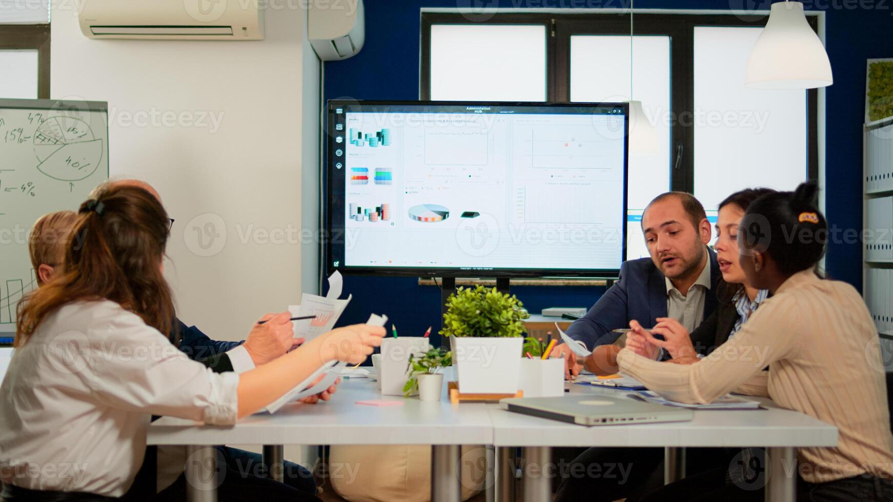 Executive explaining company's vision and potential to employees sitting at brainstorming table in broadroom with TV screen on the wall showing corporate growth photo