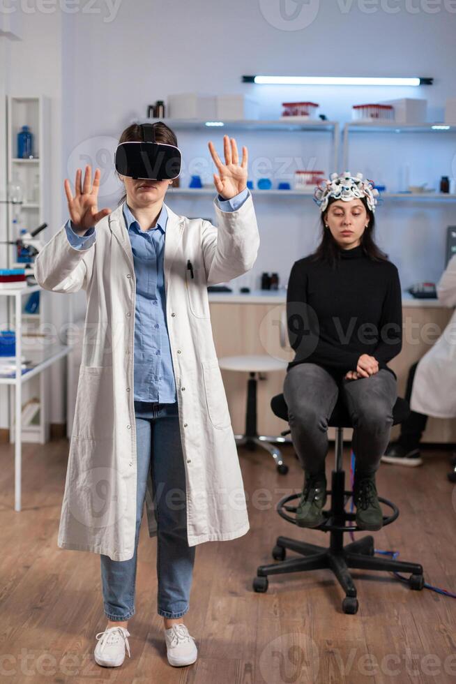 Neuroscience doctor gesturing wearing vr goggles during brain science research, patient with neurology scanner in laboratory. Doctor searching diagnosis, experiment , eeg , medicine lab. photo