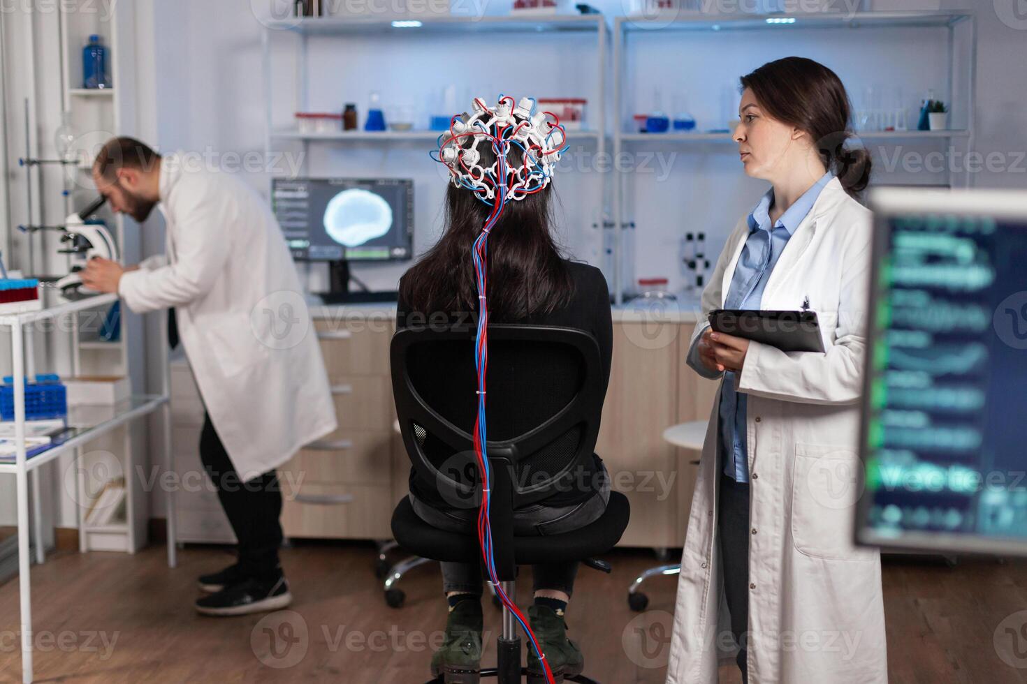Back view of woman wearing headset for brain scan in neurology laboratory, neuro science. Nervous system analysed by team of scientists. Medical scientist looking through microscope in background developing treatment. photo