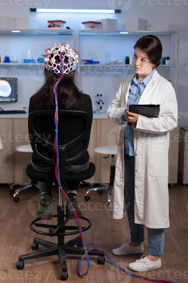 Woman patient wearing performant eeg headset scanning brain electrical activity in neurological research laboratory while medical researcher adjusting it, examining nervous system typing on tablet. photo