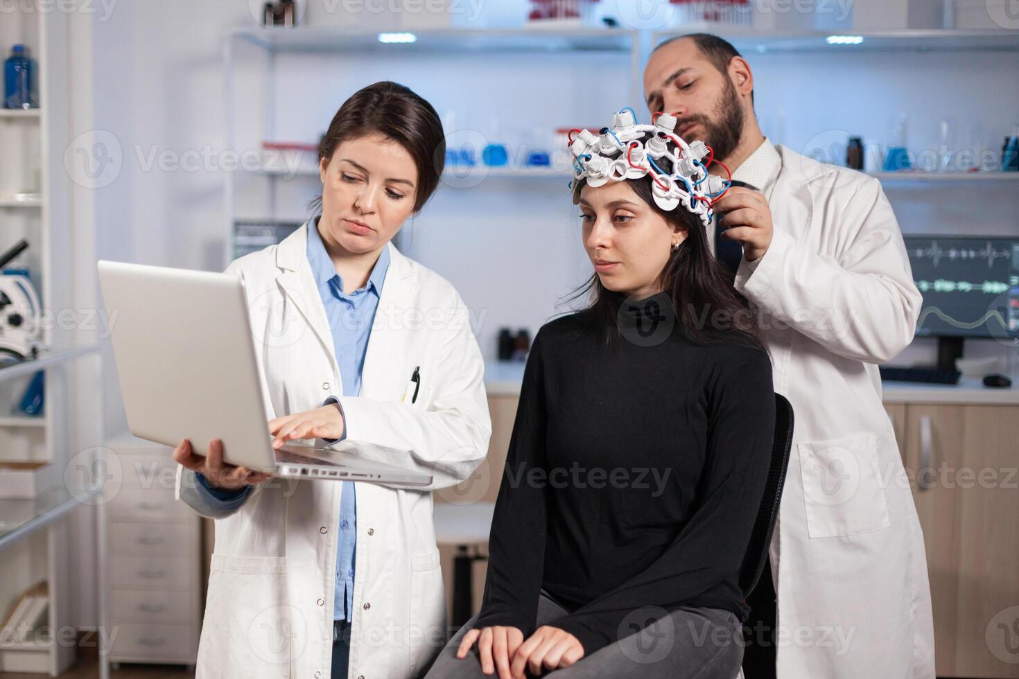 Specialist neurologist doctor taking notes on laptop asking patient's symptoms adjusting high tech eeg headset. Doctor researcher controlling EEG headset analyzing brain functions and health status. photo
