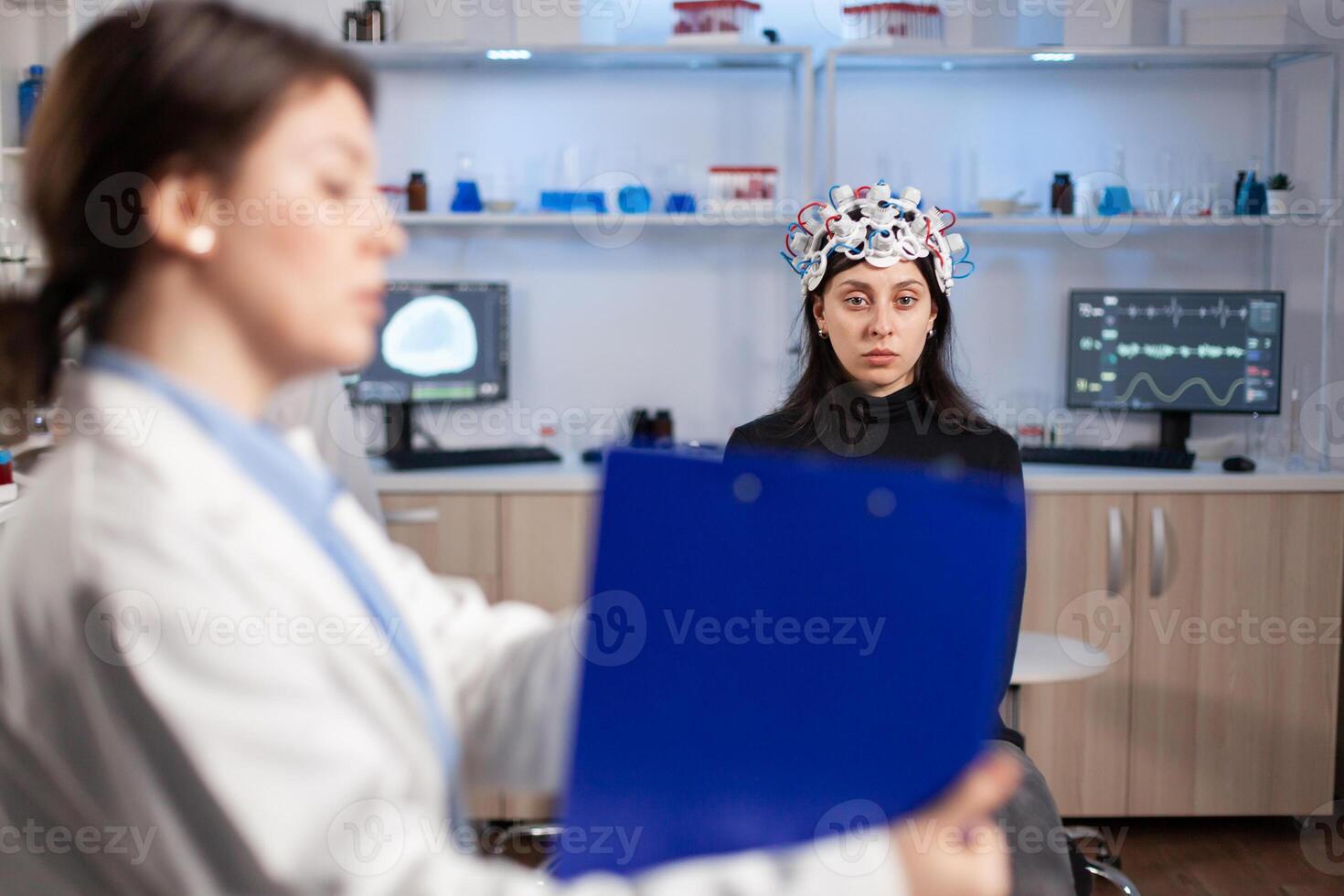 Neuroscience doctor holding clipboard showing treatment against brain disease to patient with eeg headset. Woman sitting in neurological scientific laboratory treating nervous system dysfunctions. photo