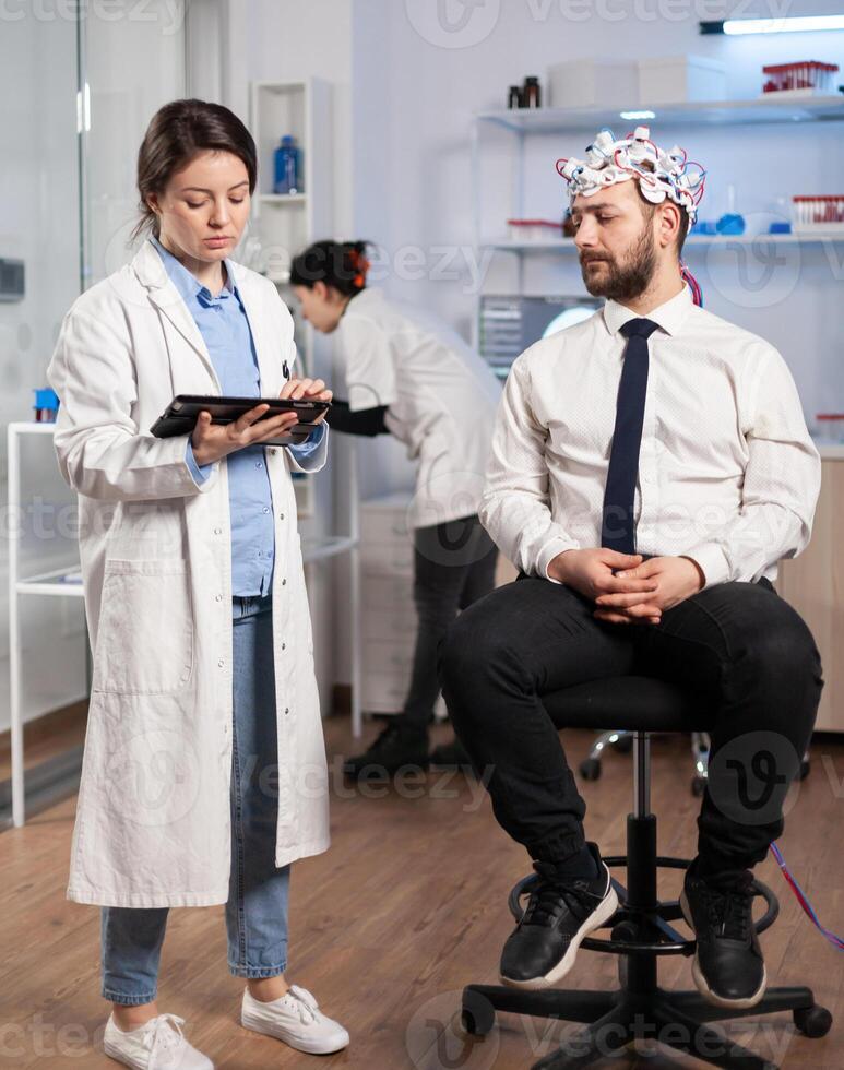 Specialist doctor in neuroscience taking notes on clipboard while testing brain functions of man using eeg headset, treating nervous system dysfunctions in moden laboratory. photo