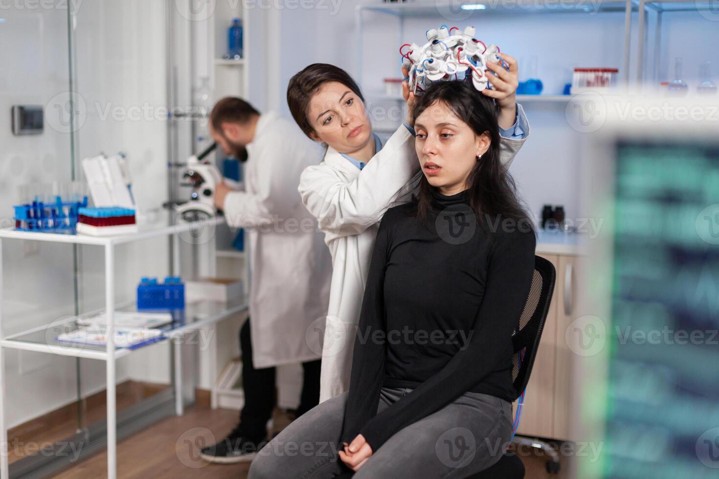 Woman wearing brainwave scanning headset sits in chair in modern laboratory brain study, neurologist looking through microscope in modern medicine lab. Team of doctors using high tech technology. photo