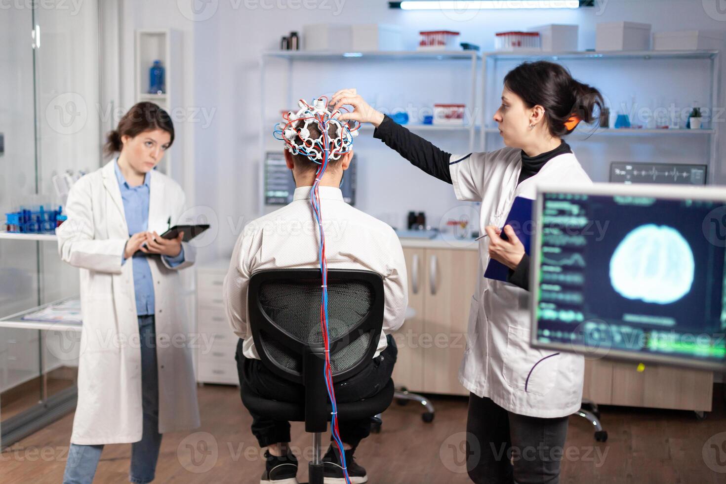 Back view of man patient wearing performant brainwave scanning headset sitting in neurological research laboratory while medical researcher adjusting it, examining nervous system typing on tablet. photo