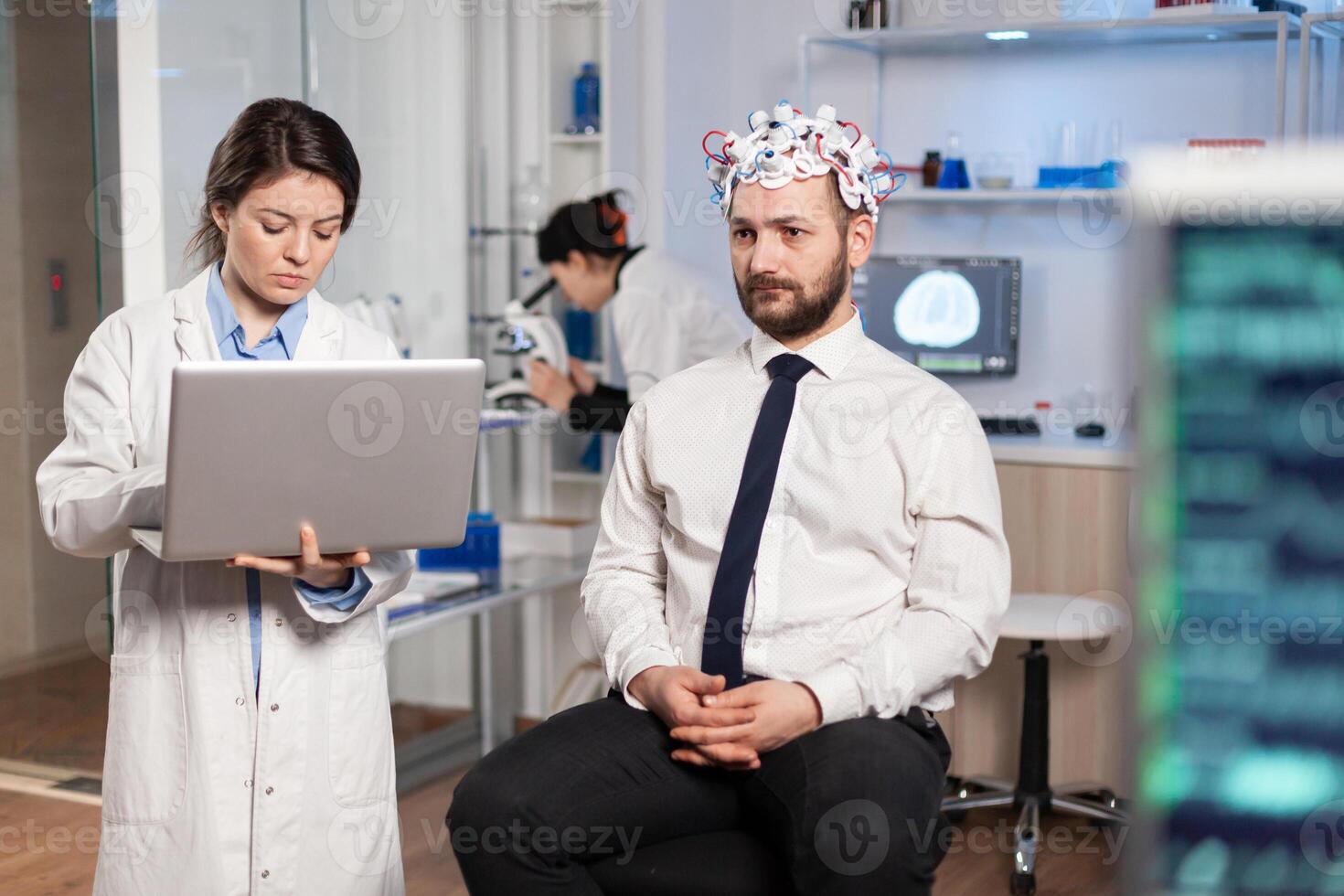 Medic in neuroscience working in neurological research laboratory developing brain experiment holding laptop explaning to man brainwave scanning headset side effects of nervous system treamtment. photo