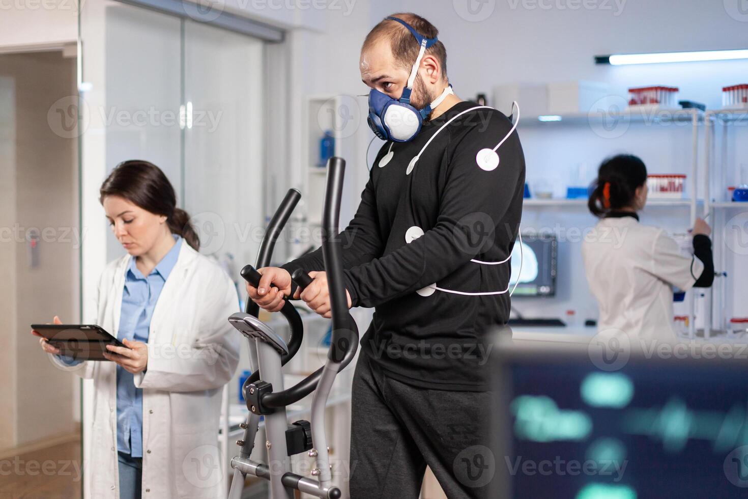 Performance athlete running on cross trainer with electrodes attached on body and mask in testing lab, scientist doctor monitoring heart rate looking at tablet pc. photo
