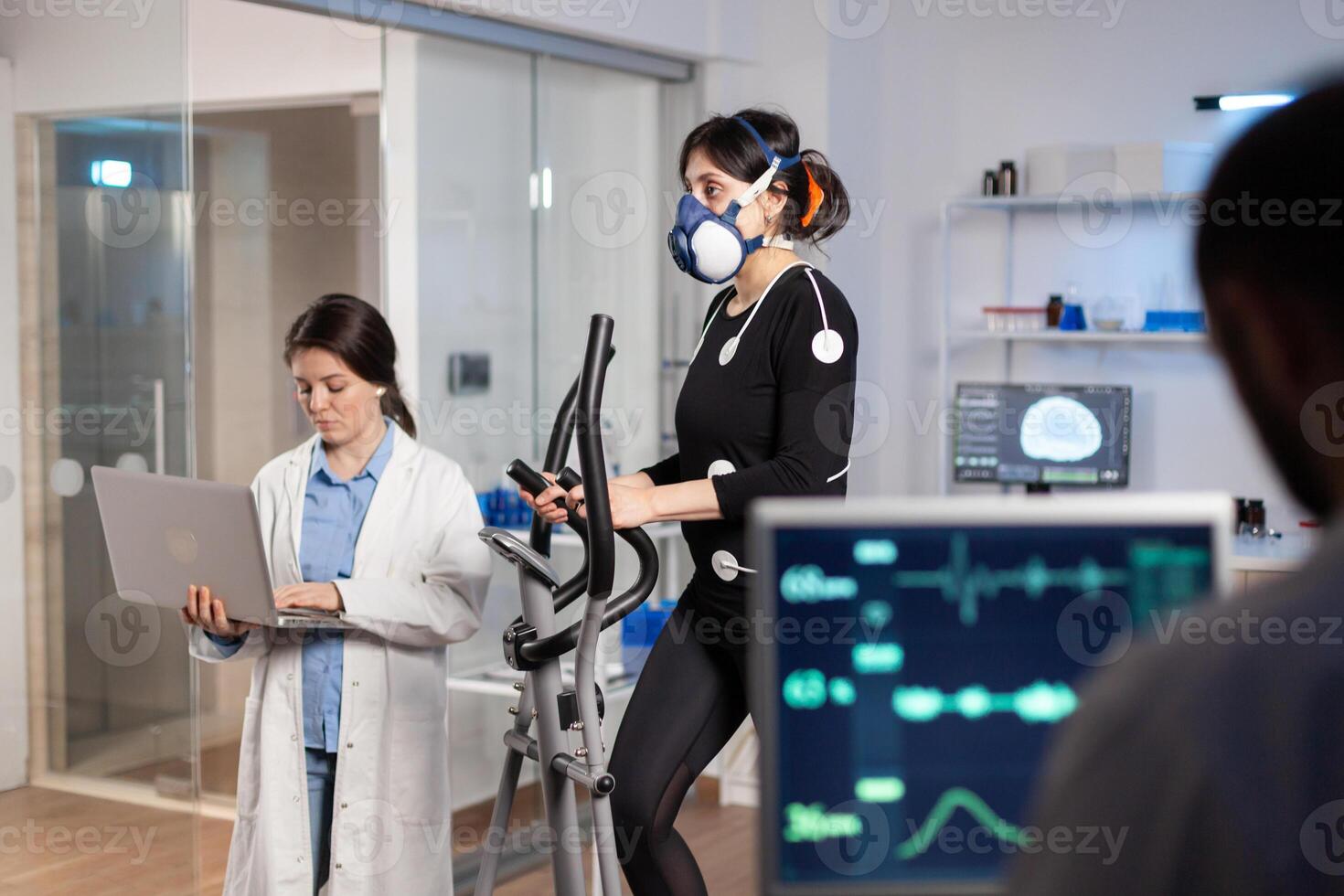 Team of medial researchers monitoring vo2 of woman performance sports wearing mask running. Lab science doctor measuring endurance using tablet while ekg scan runs on computer screen in laboratory. photo
