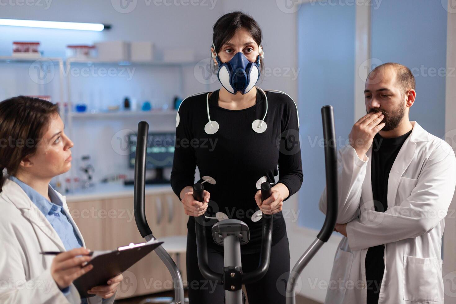 Woman athlete with mask doing fitness exercises in science sport lab with electrodes attached while scientist holding clipboard supervising whole process. Physician using notepad controling EKG data. photo