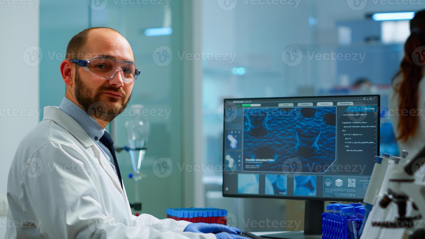 Portrait of scientist man looking at camera sitting in modern equipped laboratory. Scientist doctor examining virus evolution using high tech typing on computer chemistry tools for scientific research photo