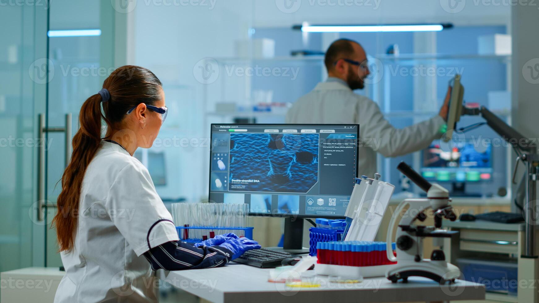 Chemistry doctor analysing blood sample and typing on computer while coworker opening door using digital computer with touch scan. Woman examining virus evolution using high tech and chemistry tools photo