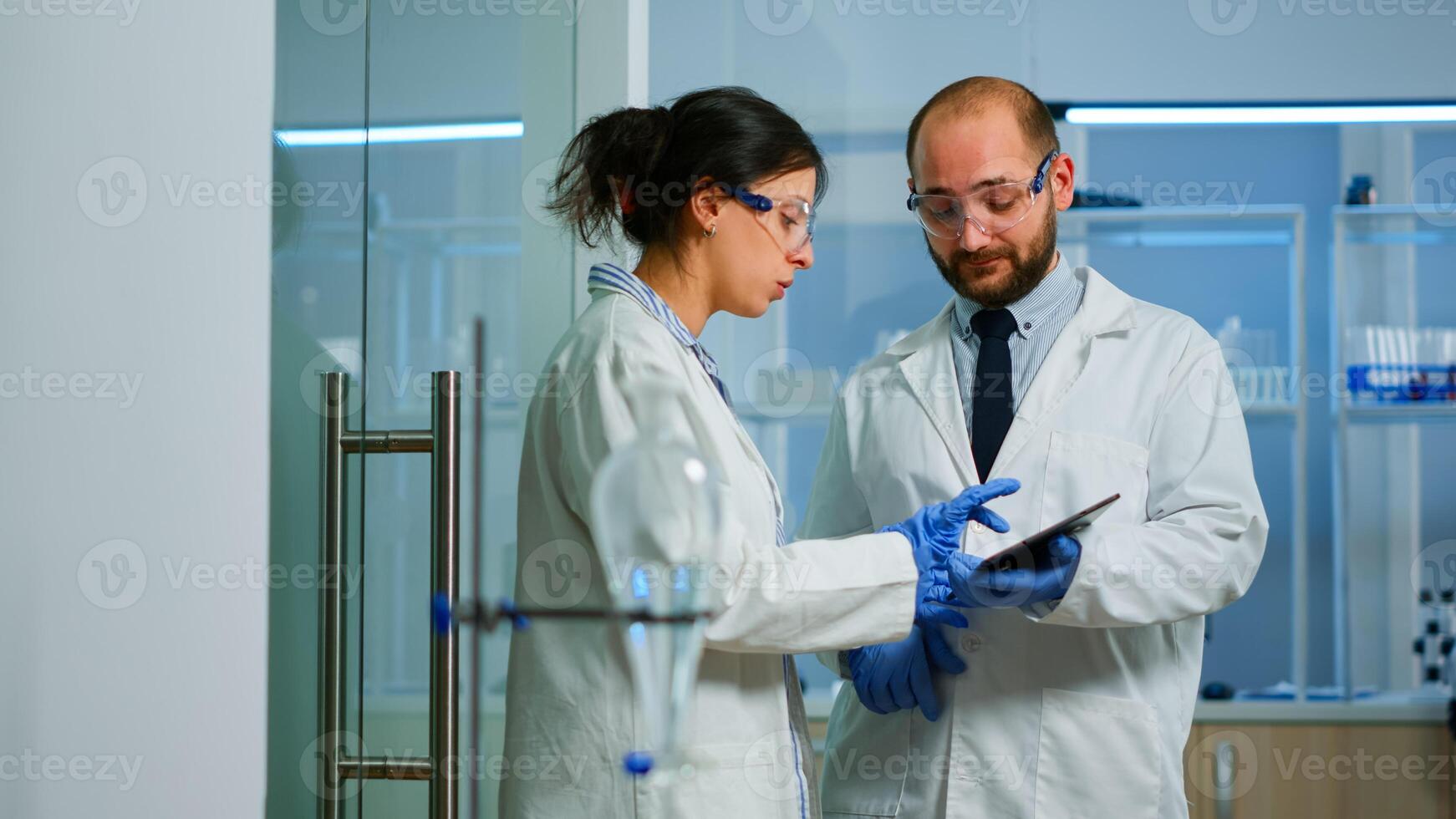 Scientist team meeting and discusssing treatment results standing in research laboratory pointing on tablet. Stuff examining vaccine development against covid19 virus using high tech for researching. photo