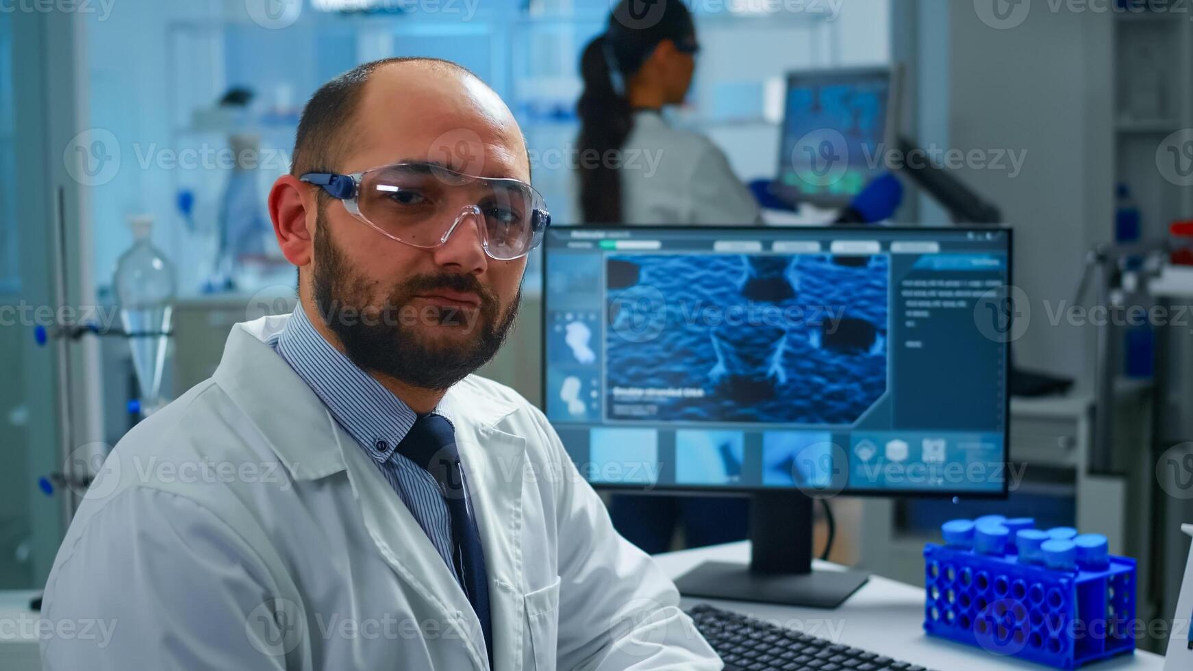 Portrait of exhausted scientist man looking at camera sitting in front of computer with DNA scan image. Chemist examining virus evolution using high tech and chemistry tools for scientific research photo