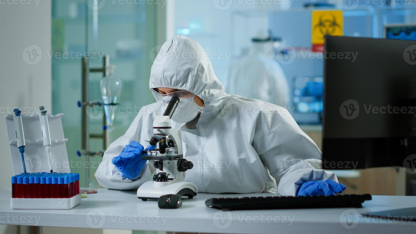 Biologist doctor in coverall checking DNA sample on microscope typing on pc in equipped lab. Examining vaccine evolution in medical lab using high tech and chemistry tools for scientific research photo
