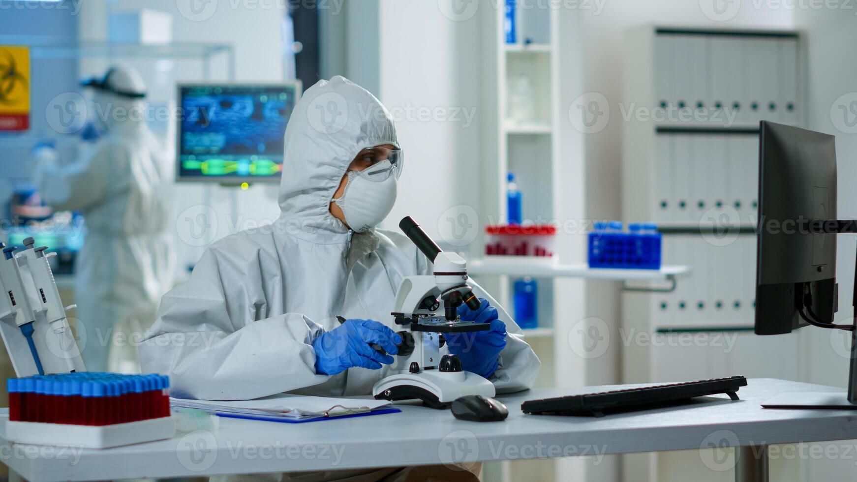 Medical research scientist conducting DNA experiments under microscope wearing protection suit and writing informations on clipboard. Chemist using high tech for vaccine development against covid19 photo