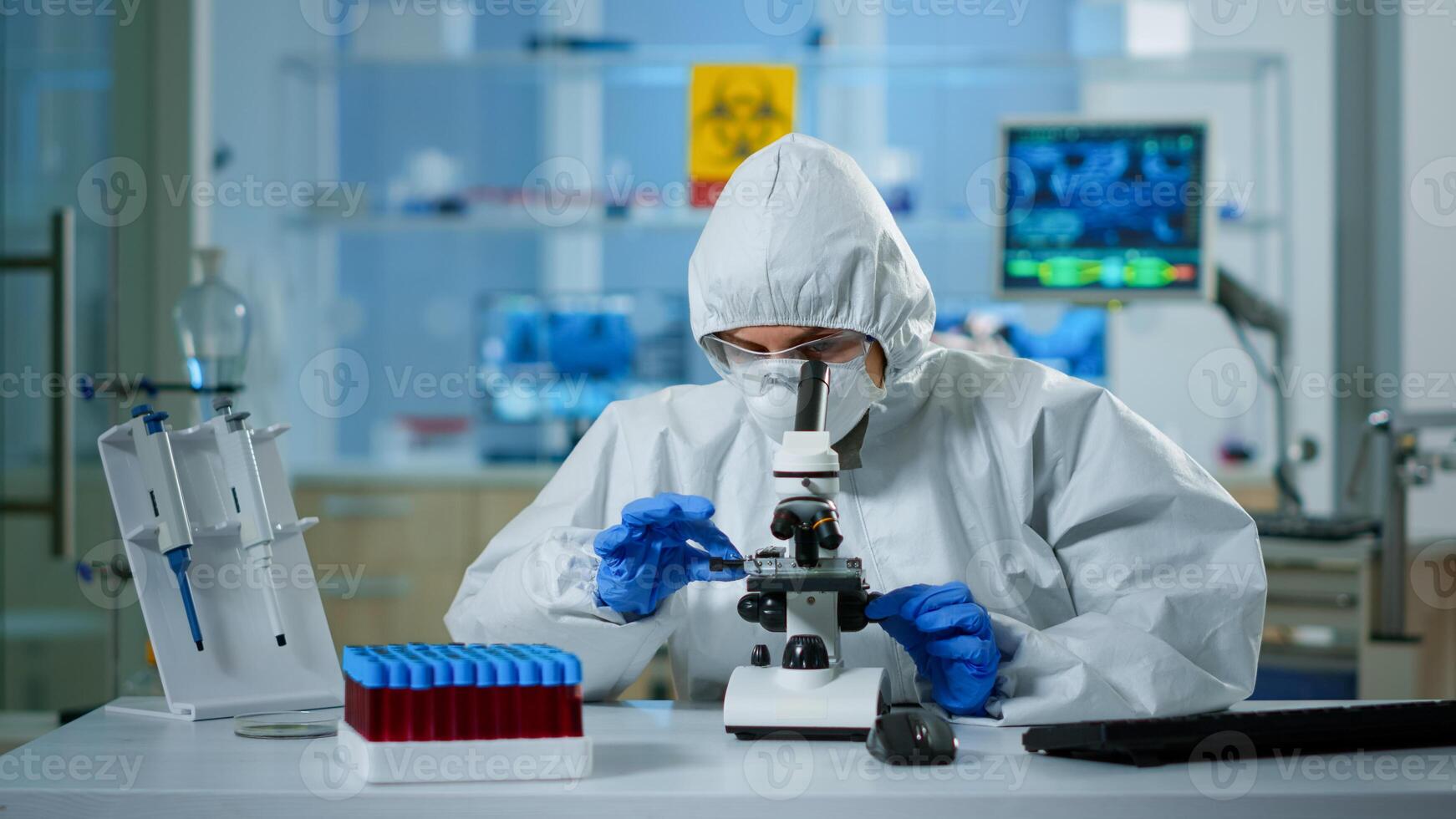 Scientist in ppe suit making adjustments and looking through laboratory microscope, typing on pc. Chemist in coverall working with various bacteria, tissue blood samples for antibiotics research photo