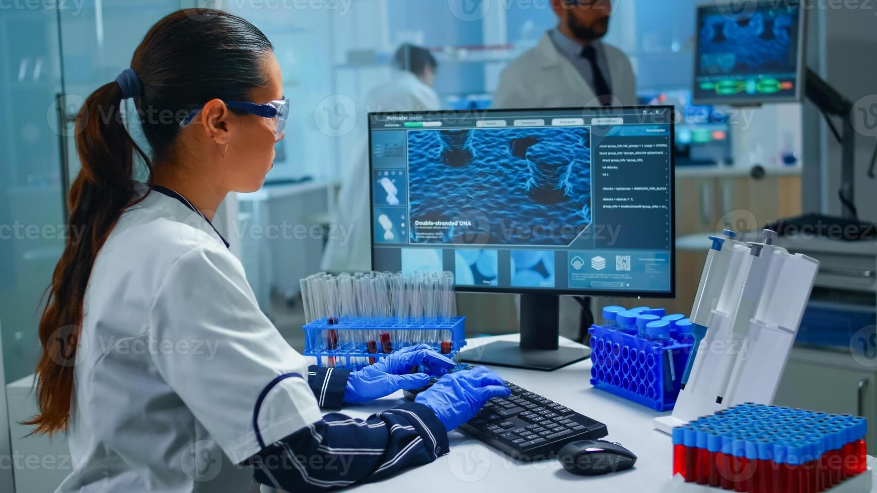 Woman technologist doing a laboratory test examining a flask with blood sample, chemist holding tube with liquids inside. Scientist working with various bacteria tissue and DNA scan image on computer photo