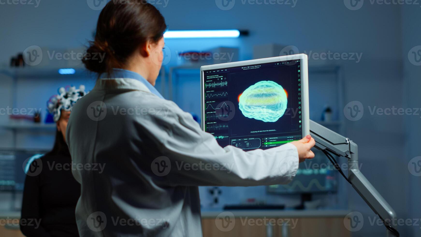 Neurologist doctor reading informations of patient looking at monitor analysing brain scan discussing with patient wearing eeg headset. Scientist taking notes examining nervous system of woman in lab photo