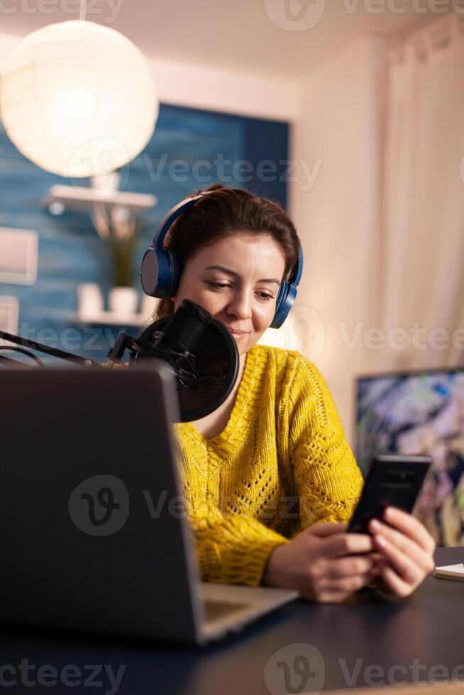 Blogger woman recording for her blog in home studio reading messages on smartphone. On-air online production internet broadcast show host streaming live content for social media. photo