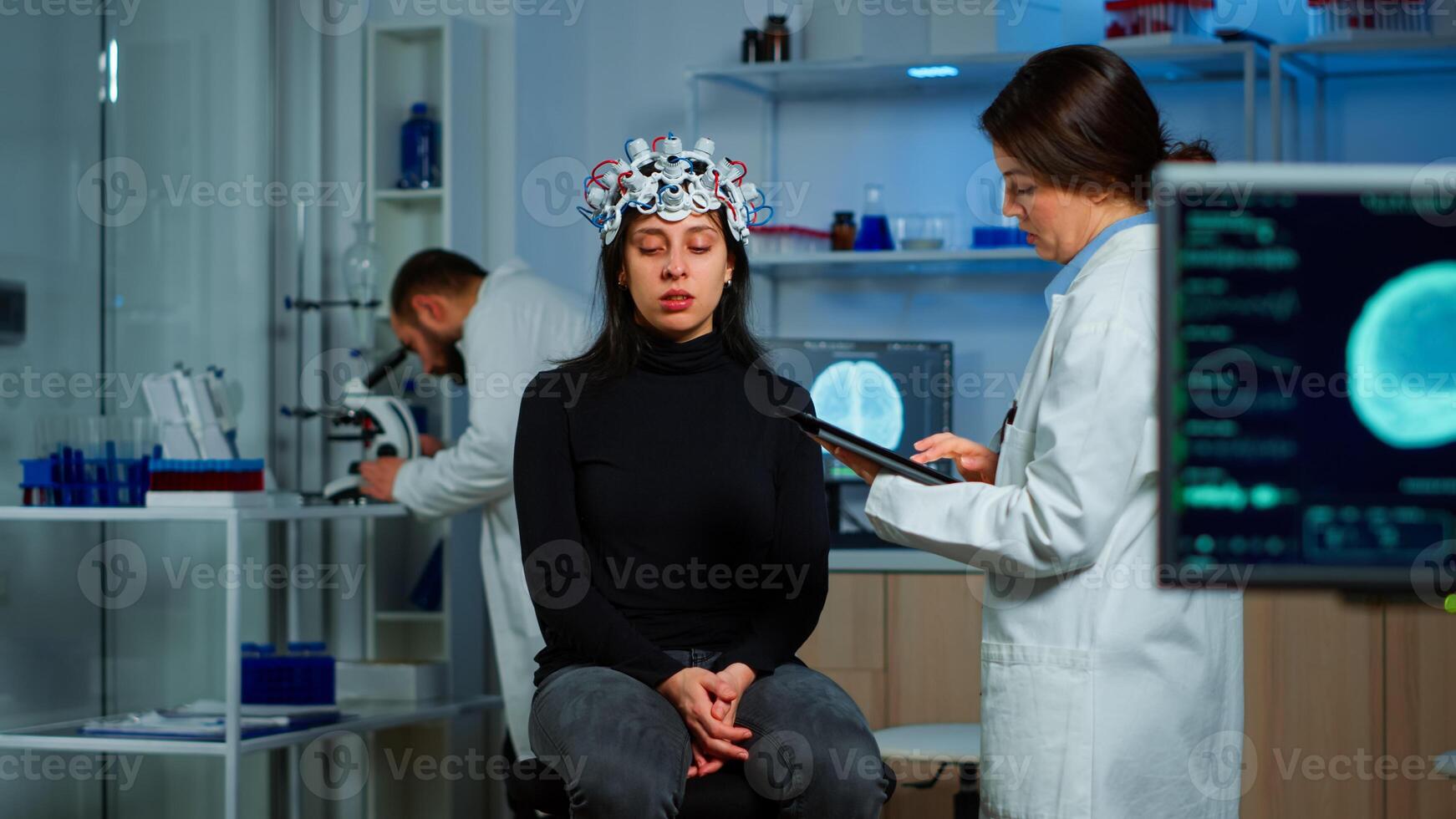 Researcher neurologist doctor asking patient's symptoms taking notes on tablet, adjusting high tech eeg headset. Doctor researcher controlling EEG headset analyzing brain functions and health status photo