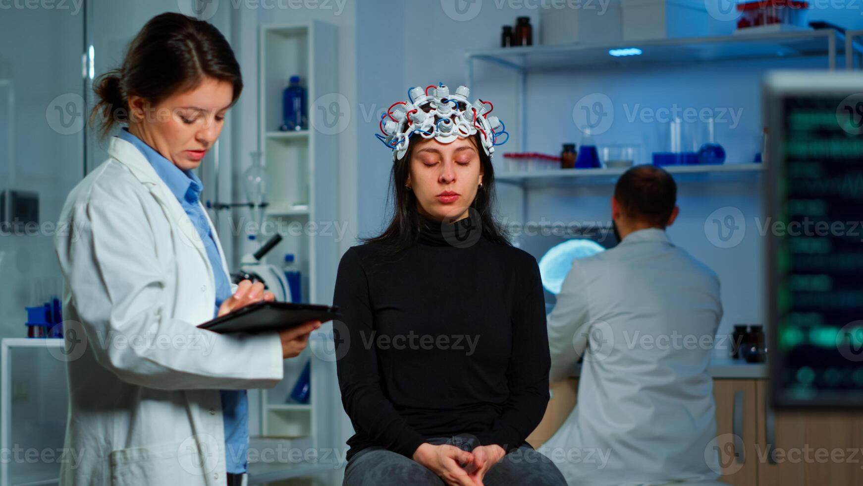 Stressed patient sitting on neurological chair with eeg headset, researcher examining health status writing on tablet. Medical scientist looking through microscope in background developing treatment photo