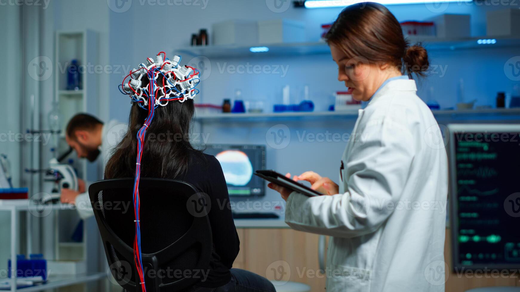 Back view of woman patient wearing performant eeg headset sitting on chair in neurological research laboratory while medical researcher adjusting it, examining nervous system typing on tablet. photo