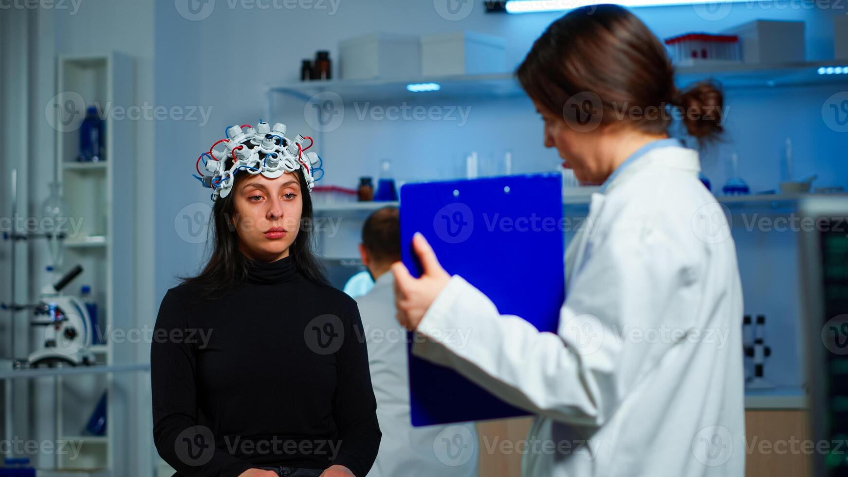 Ill woman listening neurological doctor looking at clipboard wearing high tech headset scanning brain electrical activity. Patient sitting in scientific laboratory treating nervous system dysfunctions photo