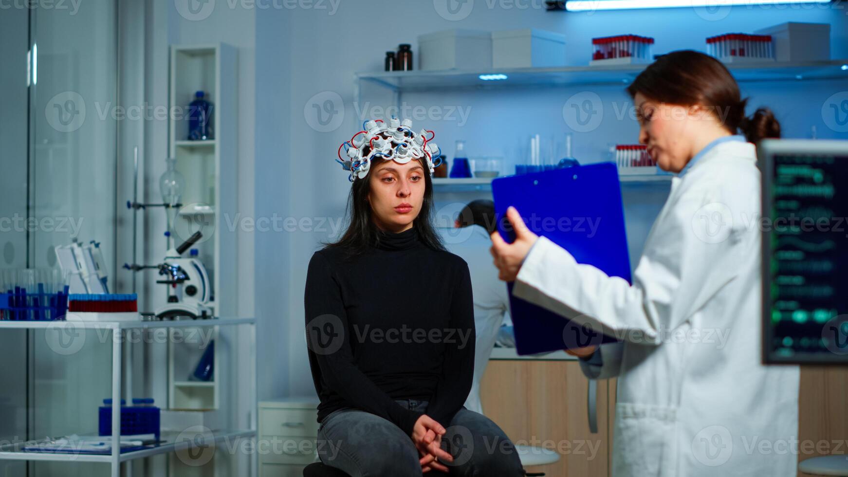 Medic researcher pointing at clipboard explaning treatment against brain disease to patient with eeg headset. Woman sitting in neurological scientific laboratory treating nervous system dysfunctions photo