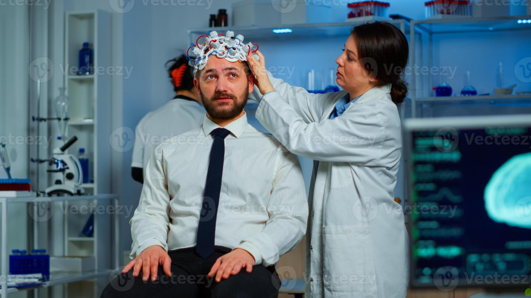 Woman researcher putting brainwave scanning headset to patient developing experiments using brain study lab monitors. Man siting on chair while scientist adjusting device examining nervous system photo