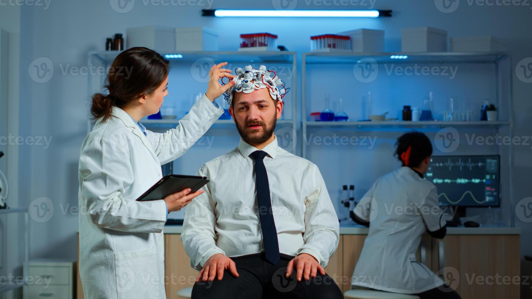 Young man wearing brainwave scanning headset discussing with neurological researcher about symptoms of disease. Medical scientist writing informations of brain scan on tablet working in laboratory photo
