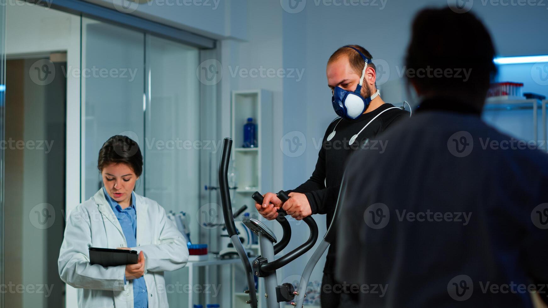 Patient making electrocardiogram during physical exercise in sports science lab, monitoring endurance and heart rate, computer showing EKG data on display. Man running with mask and medical electrodes photo