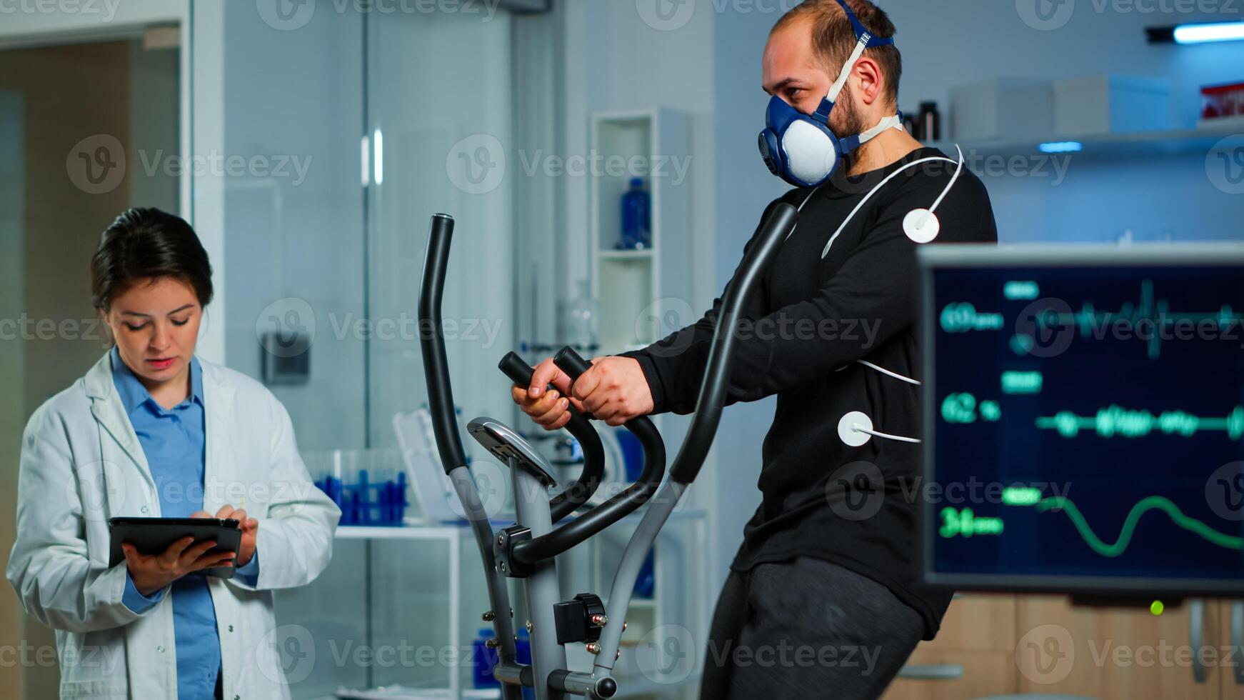 Monitor showing EKG reading of athlete running on cross trainer, while medical specialists supervise exercise, controlling physical activity, measuring heart rate in sports science laboratory photo