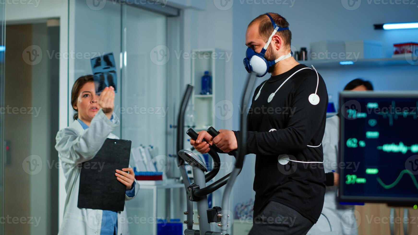 Medical doctors in sports science lab measuring performance cardiorespiratory of athlete. Team of researcher monitoring vo2 of sportsman running on cross trainer equipped with electrodes and mask photo