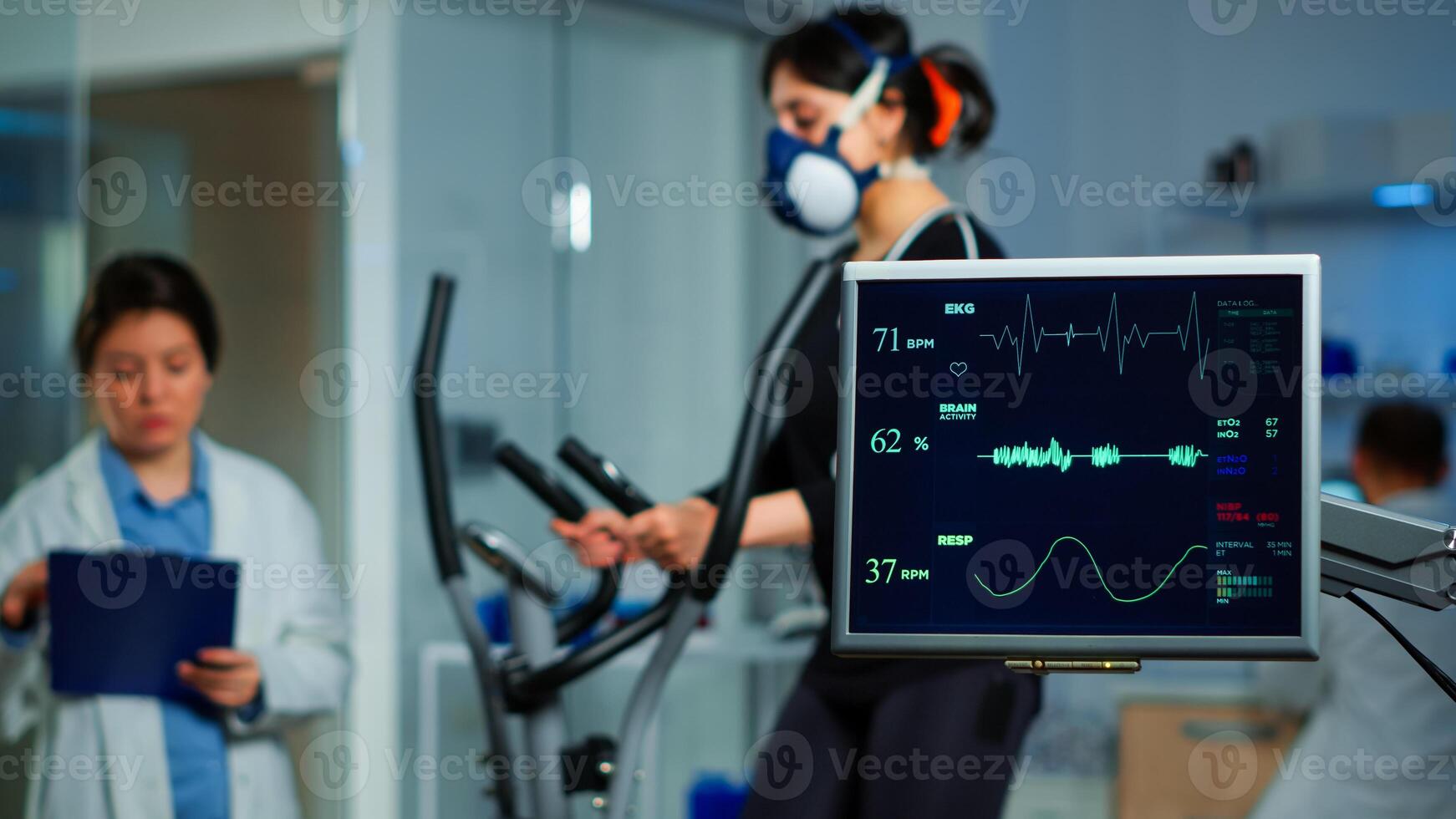 Monitor showing EKG scan of athlete running on cross trainer and medical specialists supervise exercise in background, controlling physical activity, measuring heart rate in sports science laboratory photo