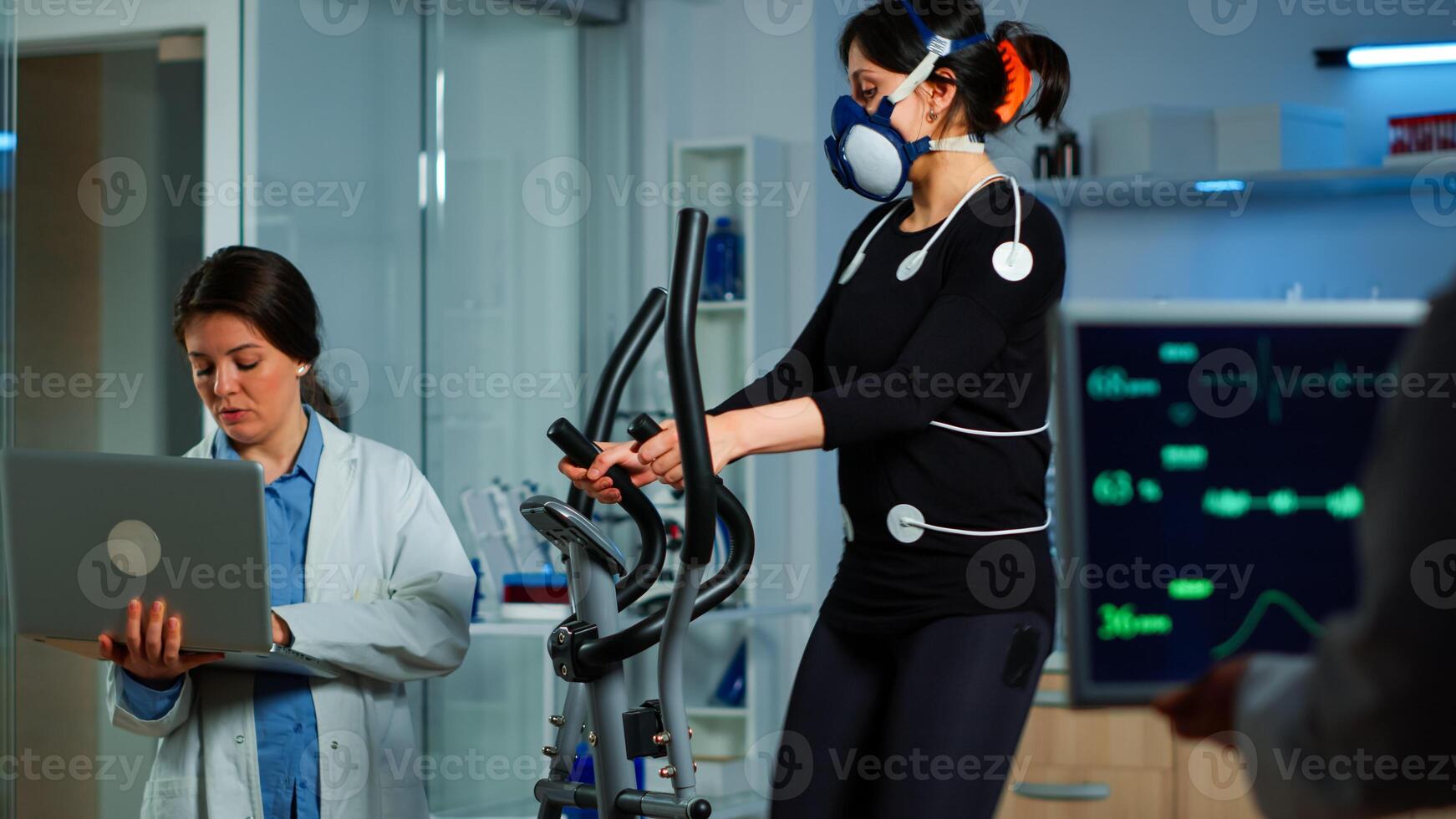 Medical researcher using laptop while measuring sportsman endurance using body sensors, electrodes and mask measuring cardiac rhythm. Patient running on cross trainer in sports science lab photo