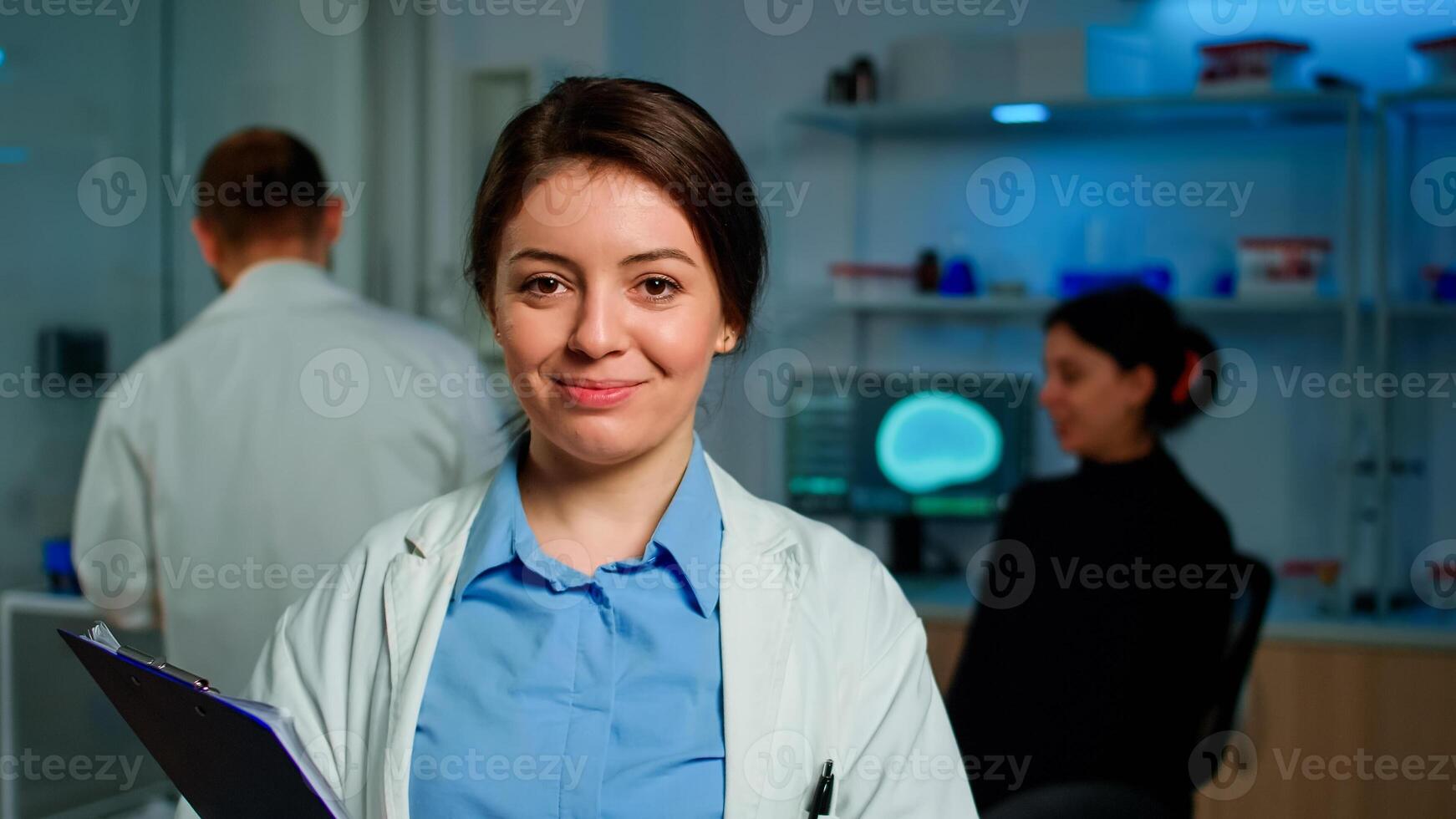 Close up of medical neurologist researcher smiling at camera standing in lab holding clipdoard while coworker discussing with patient about brain health care, nervous system, tomography scan photo