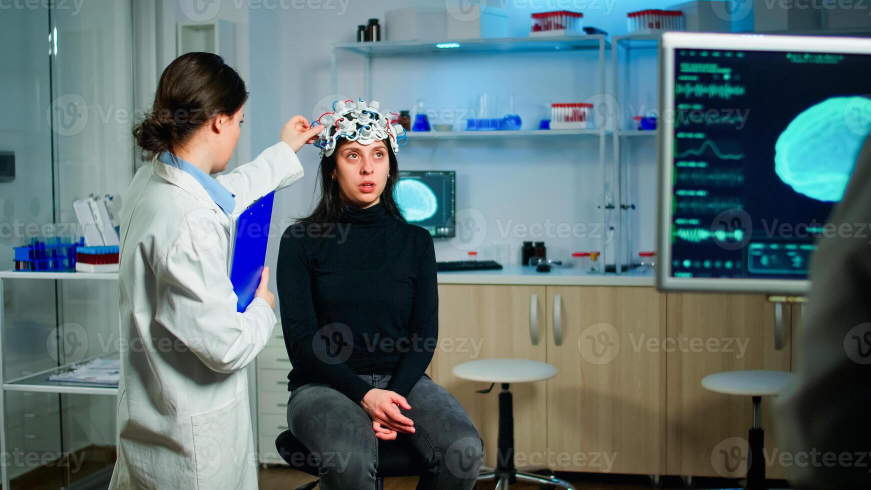 Stressed patient sitting on neurological chair with eeg headset, testing health status of brain, while medical researcher looking at monitor with tomography scan developing treatment in laboratory photo