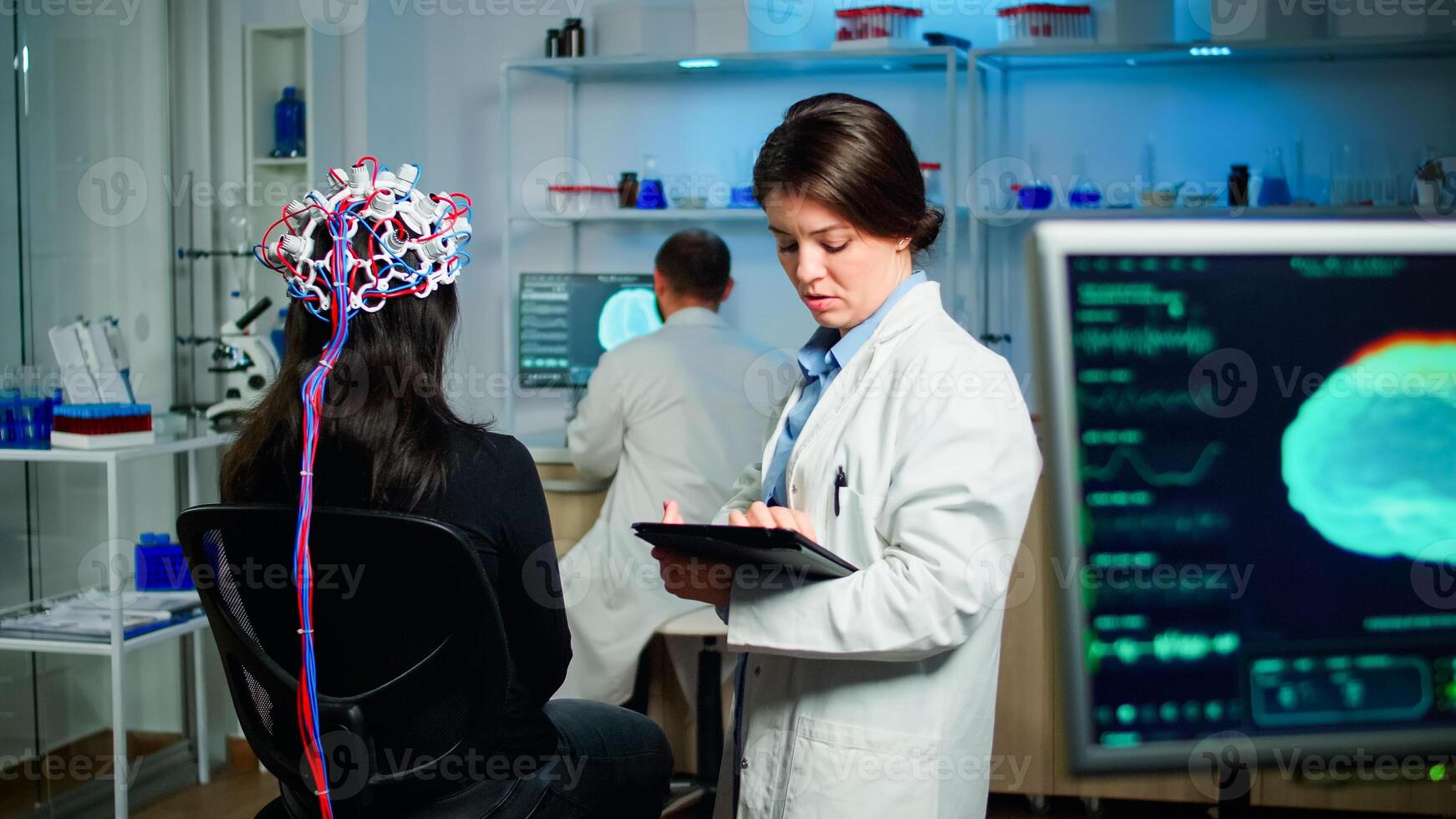 Back view of woman patient wearing performant brainwave scanning headset sitting in medical neurological laboratory while researcher adjusting it, examining nervous system typing on tablet. photo