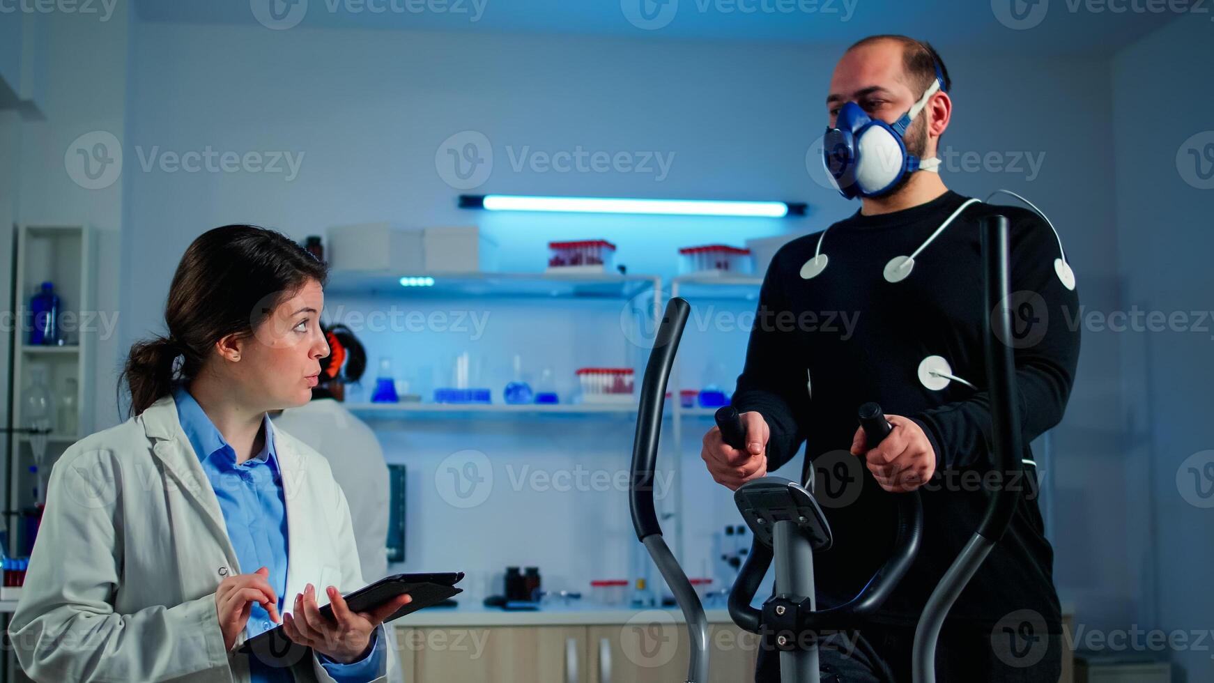 Man athlete running on cross trainer with electrodes attached to his body and mask. Physician using tablet computer and controls EKG data showing on laboratory monitors, discussing with patient photo